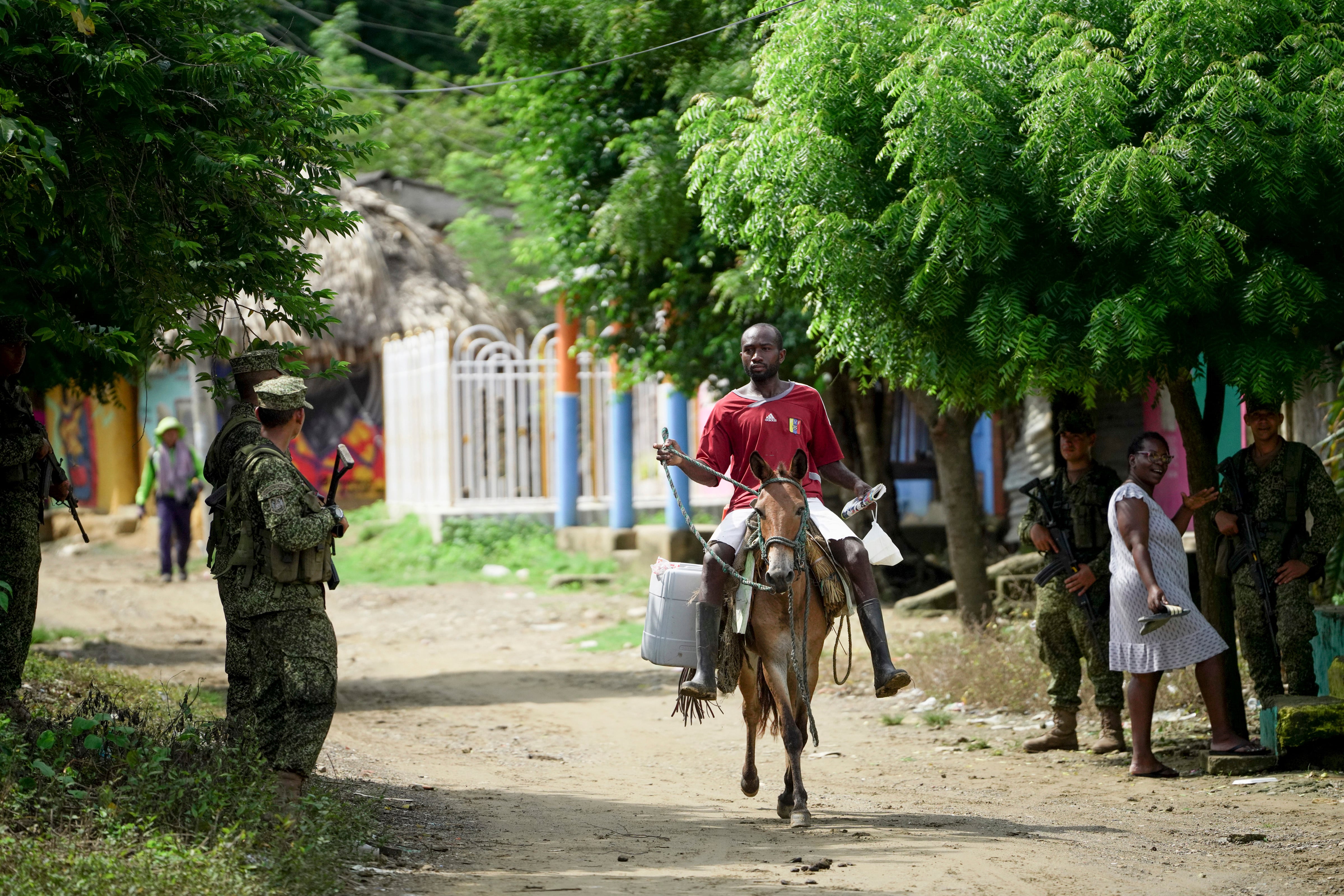 Un hombre a lomos de una mula avanza por un camino de tierra mientras residentes y soldados esperan la llegada del príncipe Enrique y Meghan, en San Basilio de Palenque, Colombia, el sábado 17 de agosto de 2024. (Foto AP/Iván Valencia)
