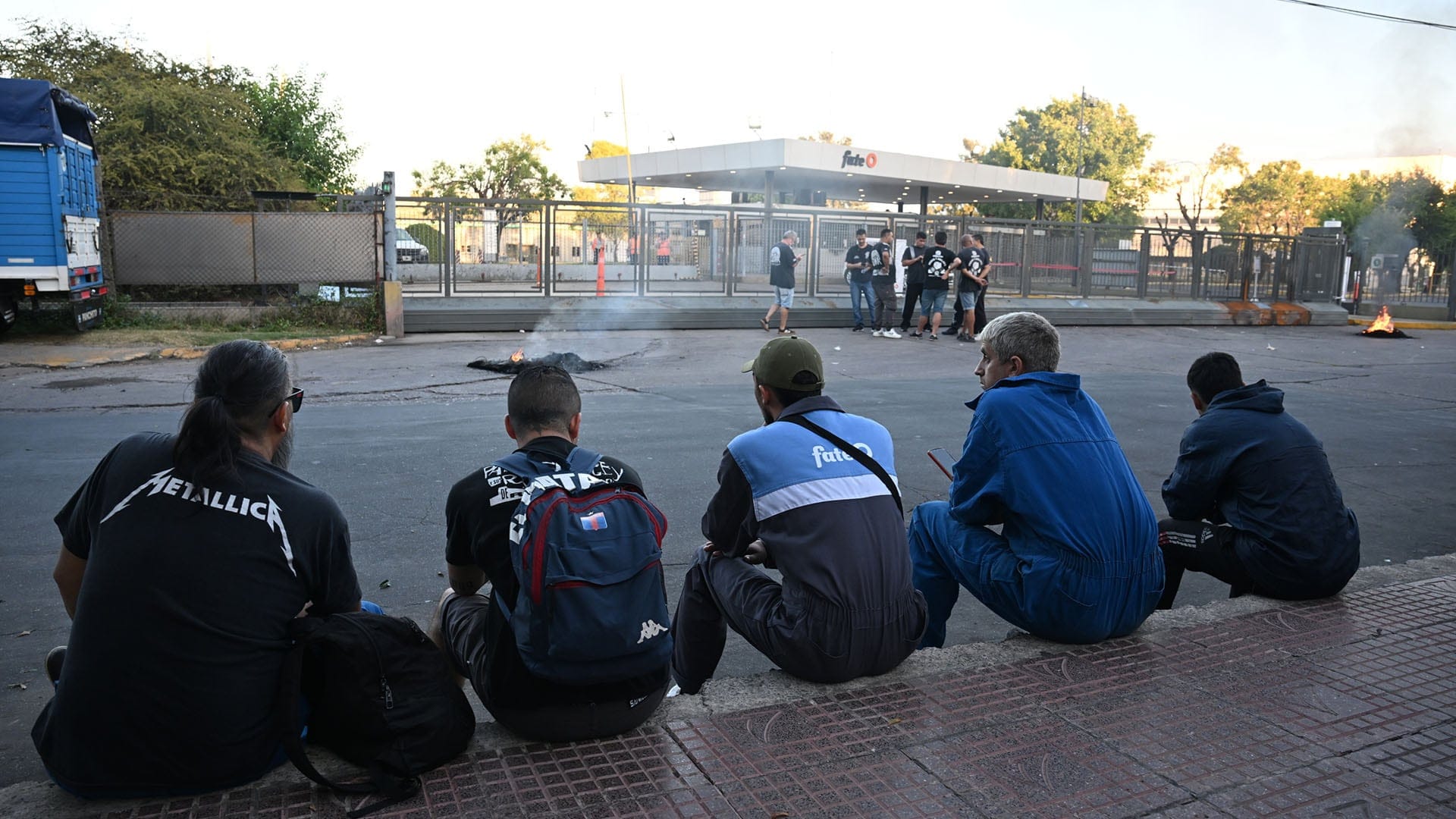 Un grupo de trabajadores se sienta en la vereda frente a la planta Fate, donde se observa un conflicto laboral, con fogatas encendidas en la calle y otros empleados congregados en la entrada. (Fotos: Maximiliano Luna)