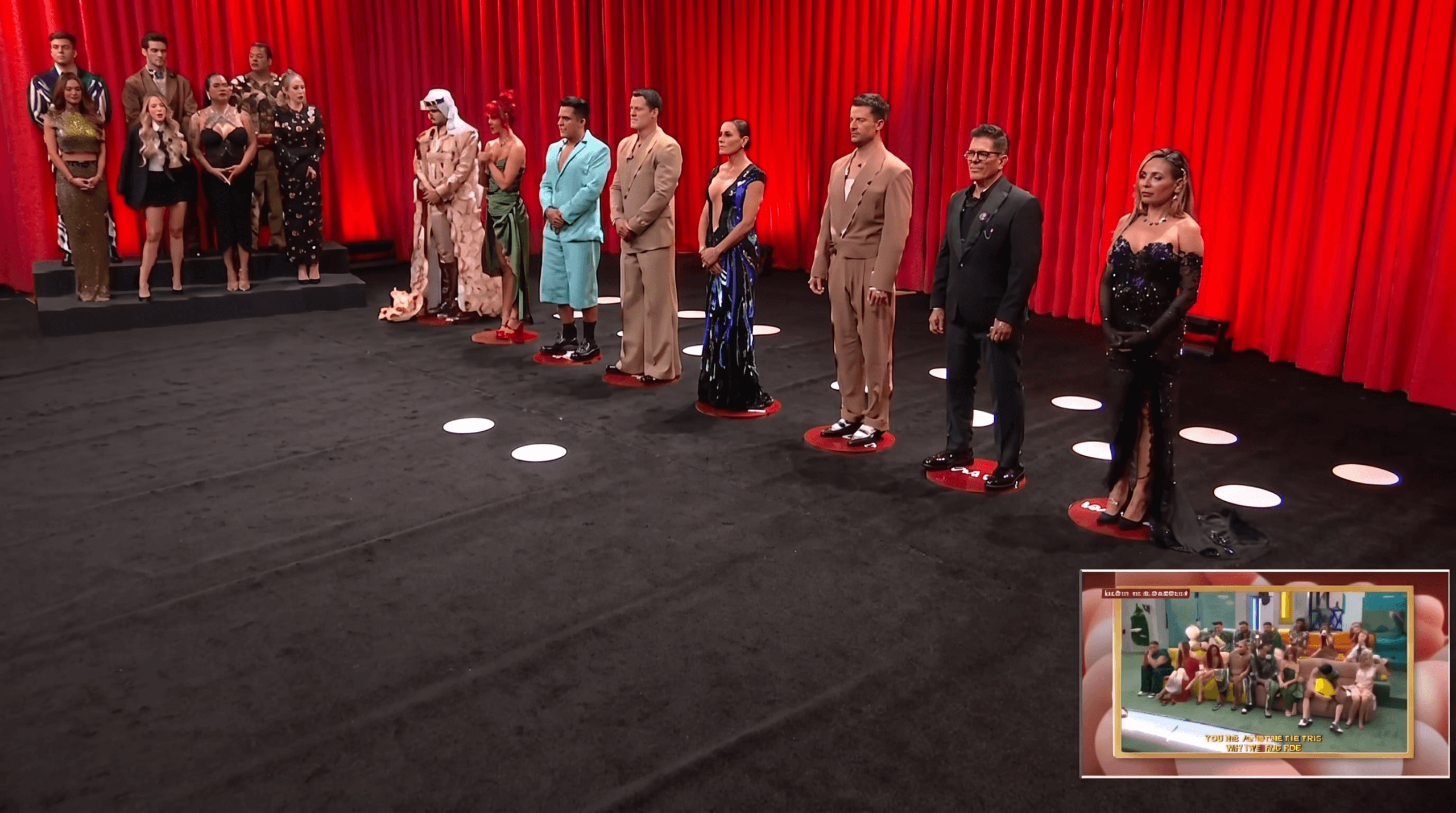 Un grupo de concursantes vestidos de gala permanece de pie sobre un escenario de televisión con fondo de cortinas rojas. (Imagen Ilustrativa Infobae)